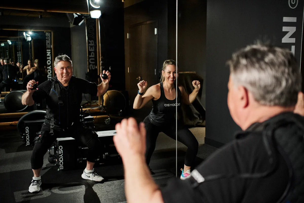 Older male member in black EMS suit doing a squat alongside a female coach in BODY20-branded top, both smiling, both moving