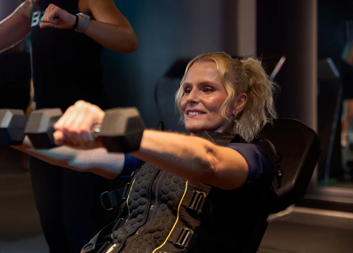 Older female member in navy EMS suit on incline chair, arms extended with dumbbells, smiling broadly, warm and capable. Coach partially visible at frame edge.